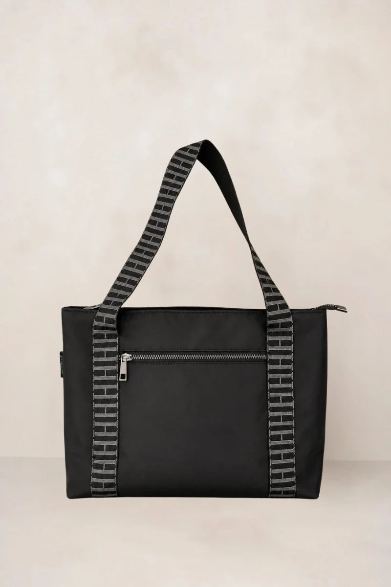 A black tote bag with a front zippered pocket and wide shoulder straps featuring a gray and black geometric pattern, set against a neutral beige background.