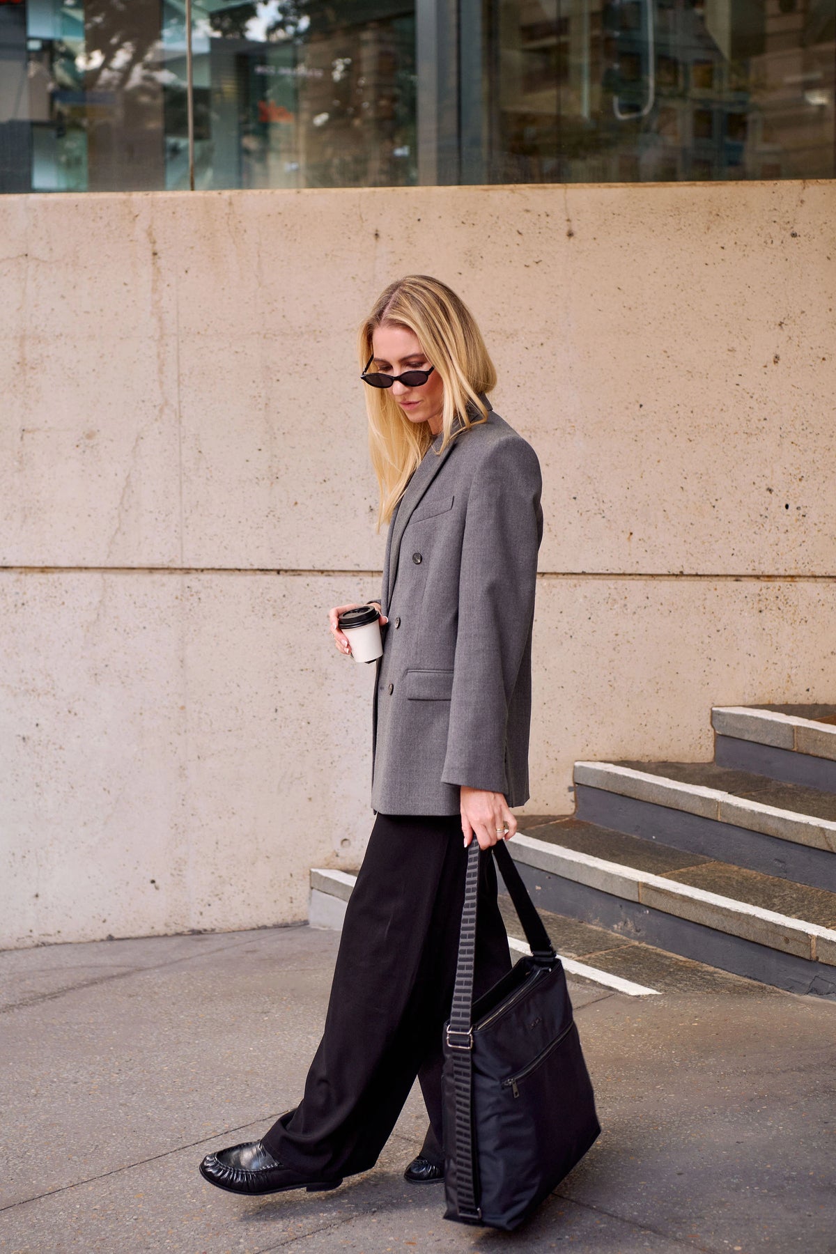 A woman walking outdoors wearing an oversized gray blazer, wide-leg black pants, black shoes, and black sunglasses, carrying a black tote bag and holding a takeaway coffee cup.