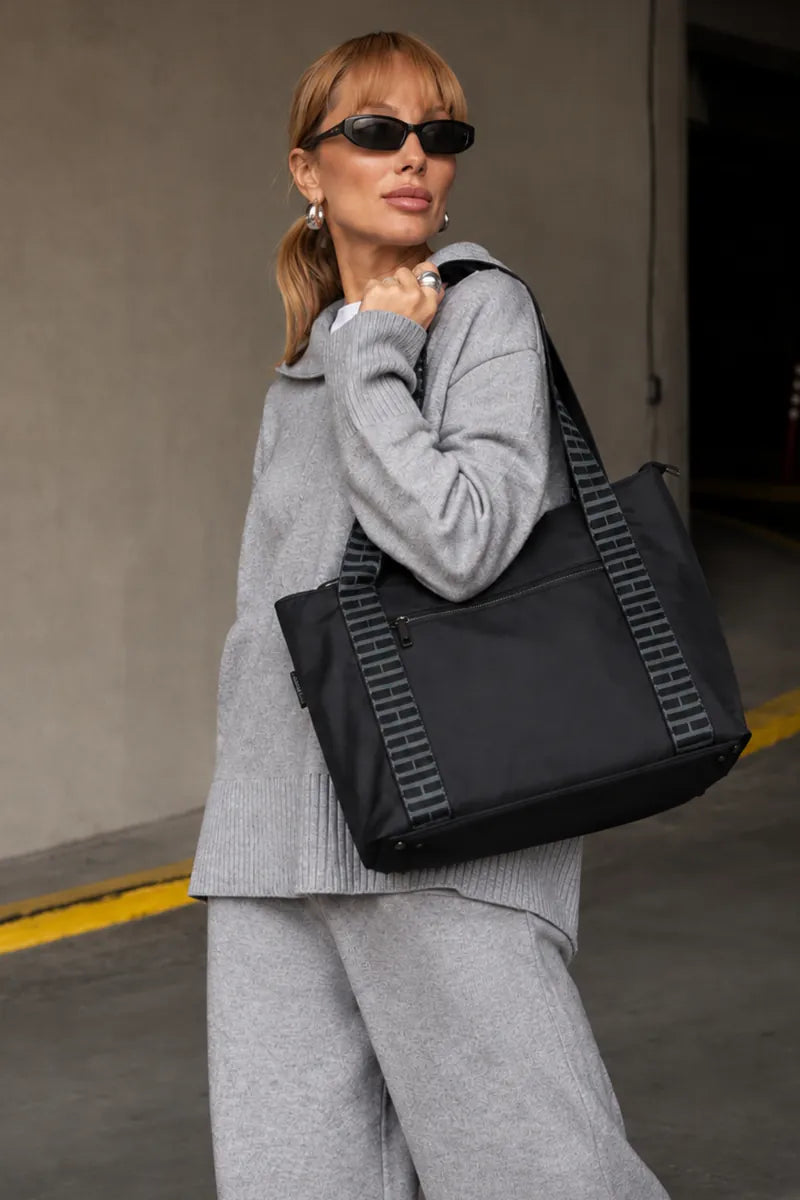 A woman in a gray knit lounge set and black sunglasses carries a large black tote bag with gray striped handles in an indoor parking structure.