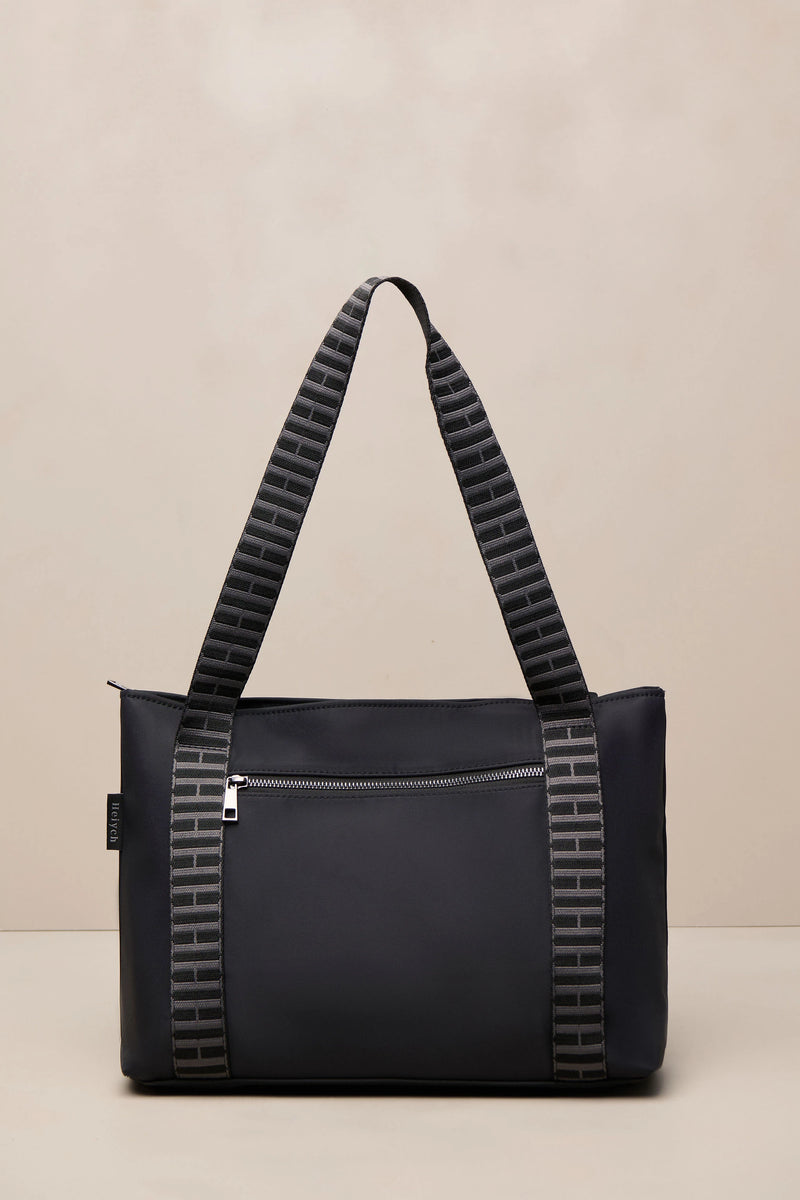 A black tote bag with textured gray and black patterned shoulder straps, shown against a neutral beige background.