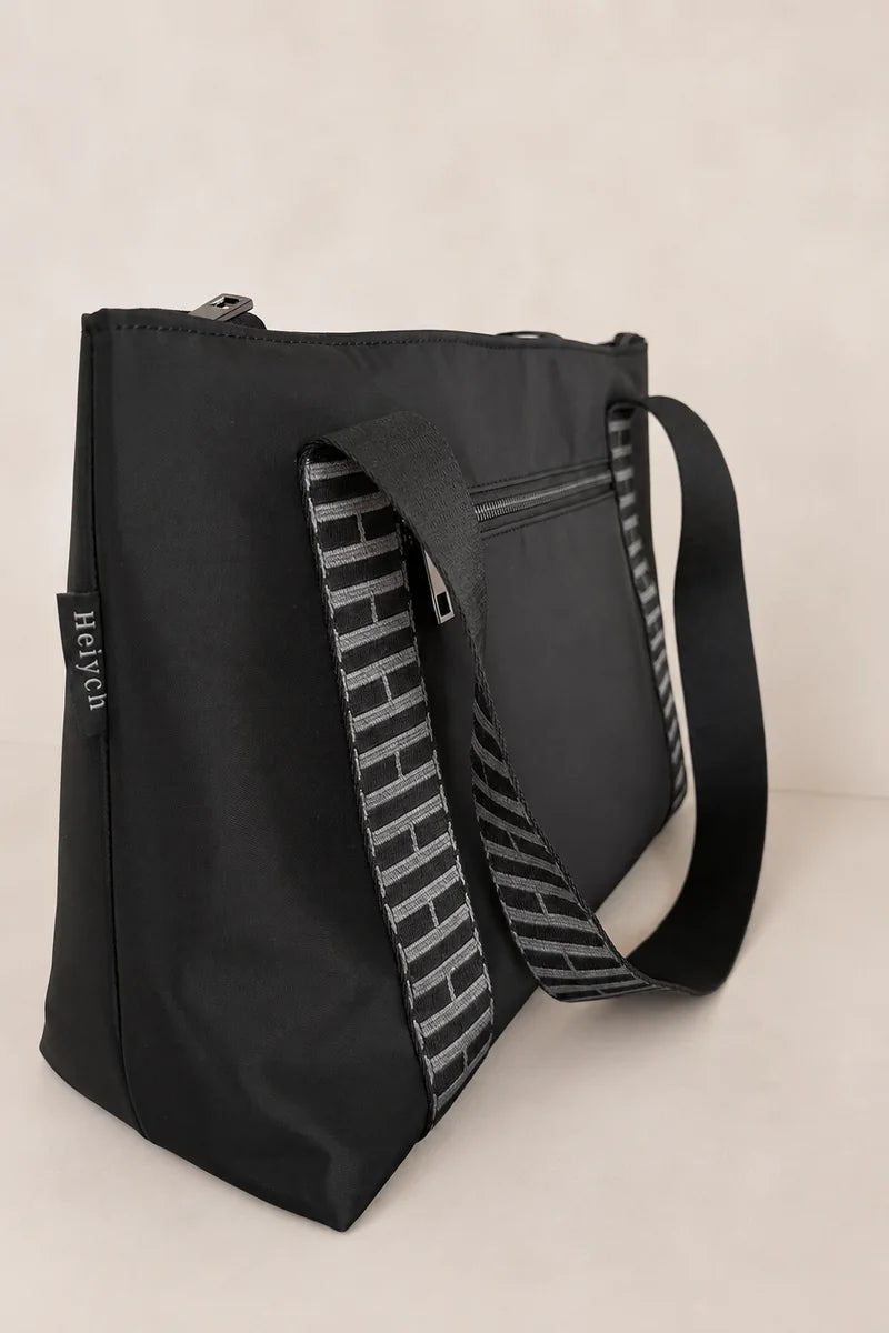 A black tote bag with a patterned gray and white shoulder strap, photographed against a light beige background.