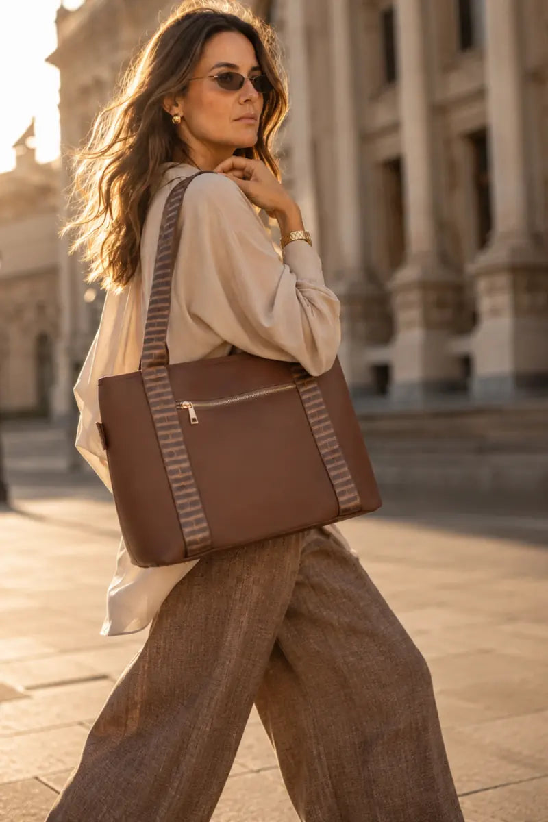 A woman walking outdoors carrying a large brown chocolate tote bag with striped fabric shoulder straps, photographed in warm golden-hour light against a blurred classical building background.