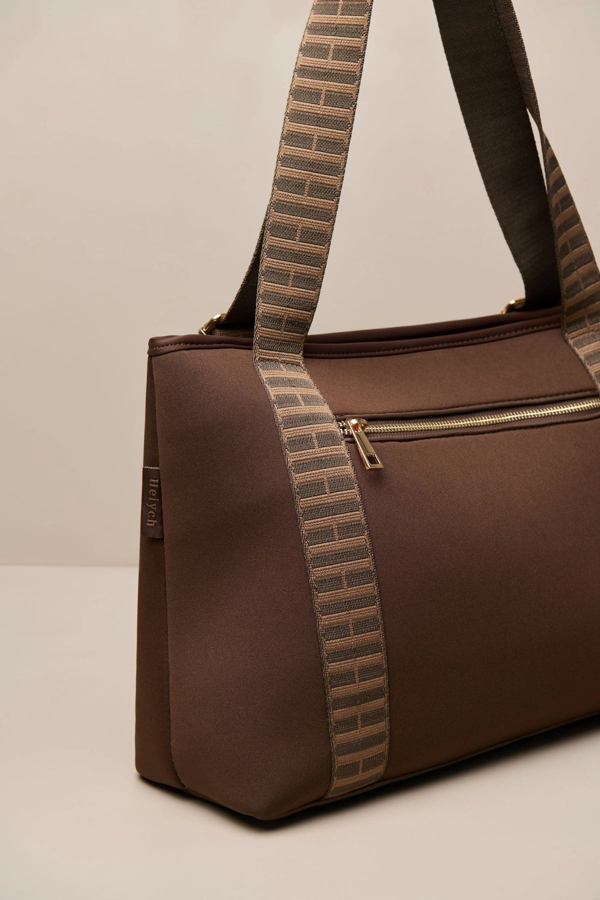 A brown chocolate duffel bag with a wide, patterned brown and beige woven shoulder strap, shown on a neutral light beige background.
