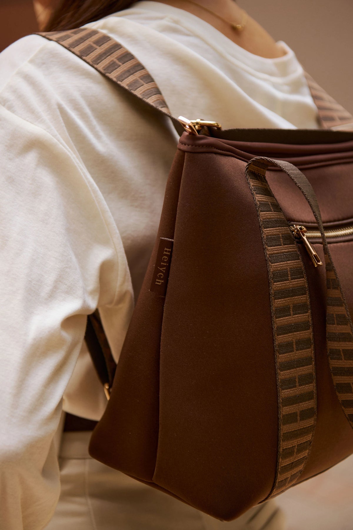 A woman wearing a white top carries a brown chocolate backpack bag with patterned brown and beige woven straps and gold-tone hardware.