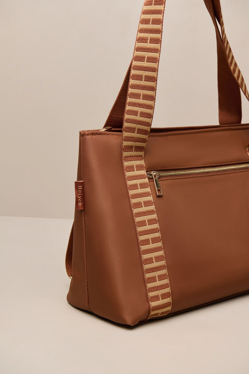 A brown cognac tote bag with a distinctive woven shoulder strap featuring a brick pattern in beige and brown, set against a neutral light beige background.