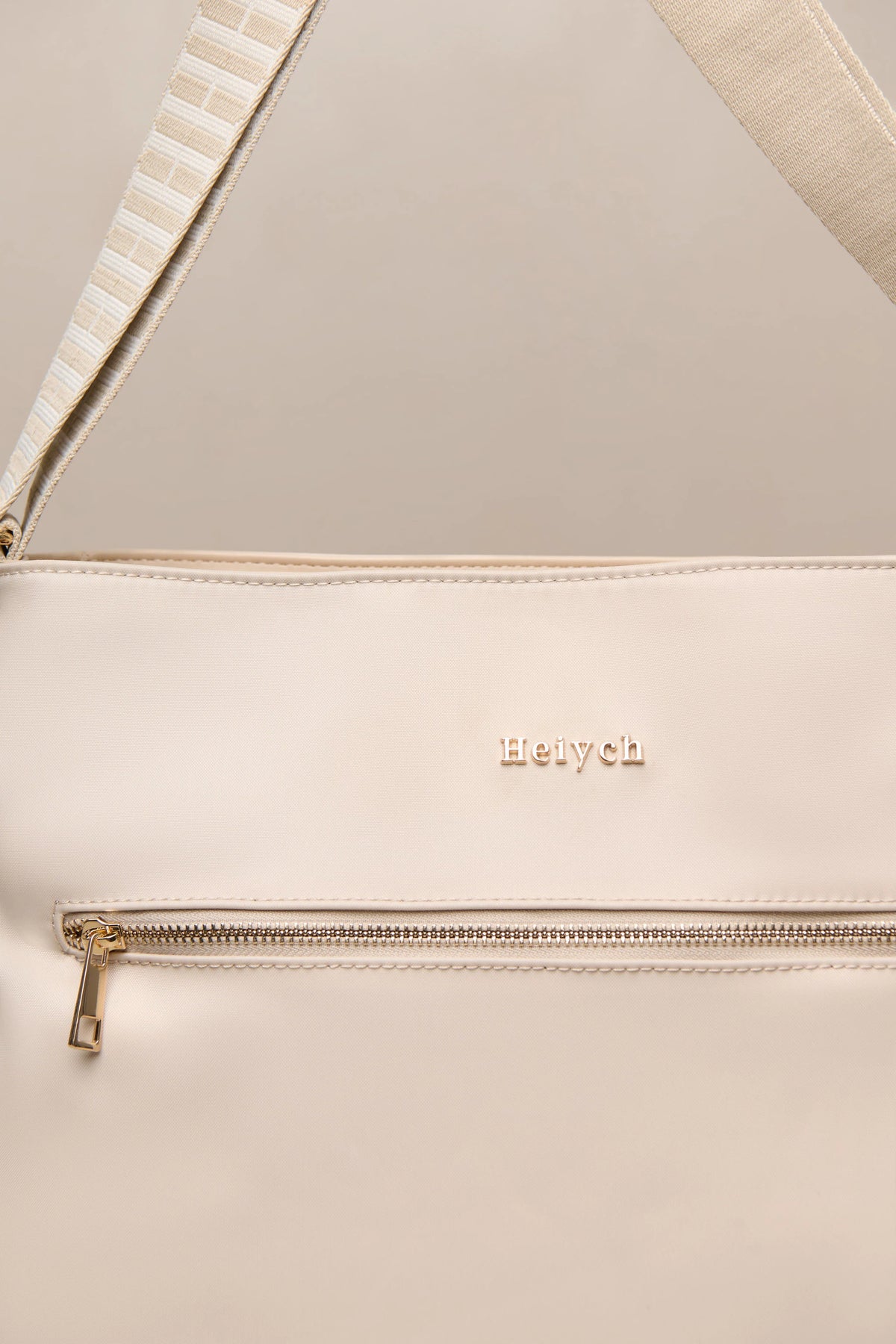 Close-up of a taupe-colored leather shoulder bag with a gold-tone zipper and the brand name 'Heiych' embossed on the front, featuring a striped woven strap detail.