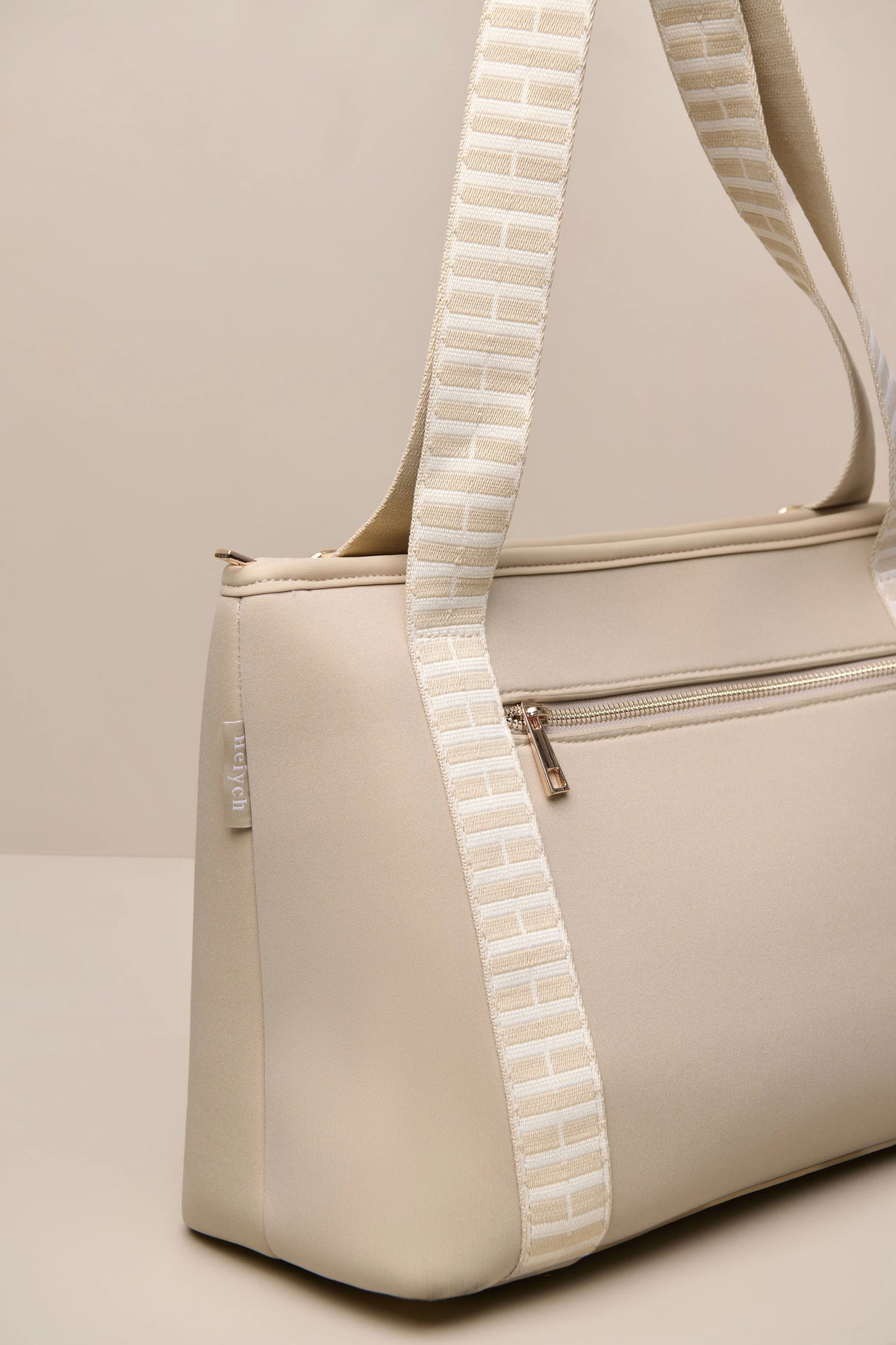 A taupe cream-colored neoprene shoulder bag with a distinctive textured, striped woven strap in a light beige and white pattern, shown against a neutral beige background.