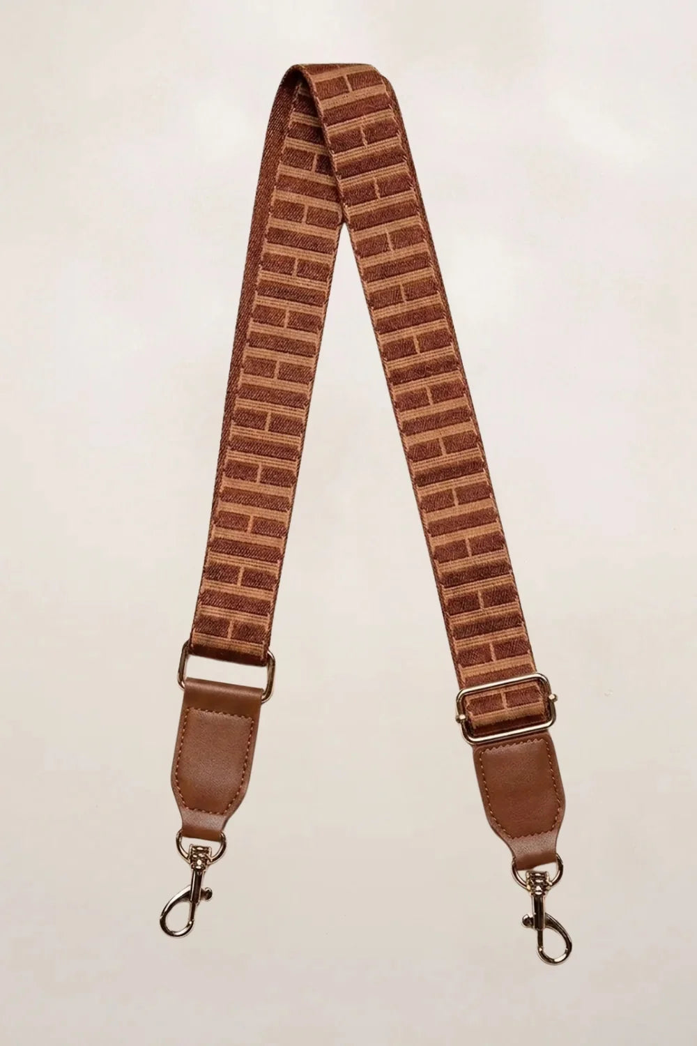 A tan fabric shoulder strap with a brick-patterned design and brown leather-like ends, featuring metal swivel clips on a light beige background.
