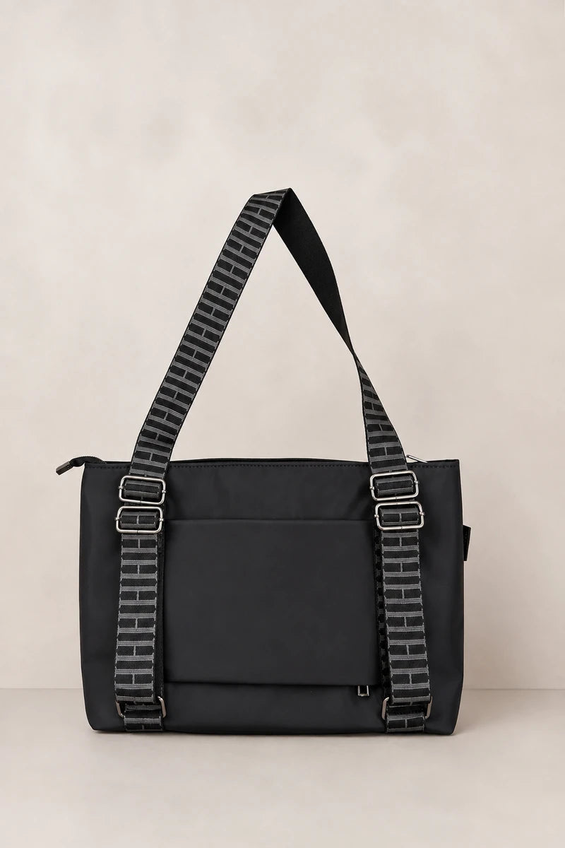 A black nylon shoulder bag with a front pocket and two wide, patterned black-and-white straps with silver buckle closures, set against a neutral beige background.