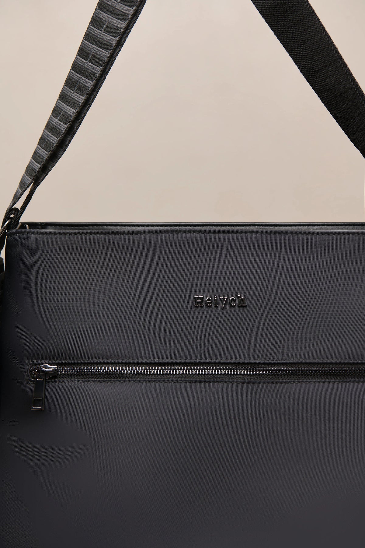 Close-up of a black nylon shoulder bag with a horizontal zippered pocket and a black-and-gray patterned strap, featuring the embossed brand name 'Heiych' on the front.