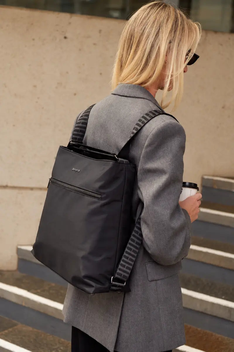 A woman in a gray blazer wearing a black nylon crossbody backpack with patterned shoulder straps, standing on outdoor stone steps.