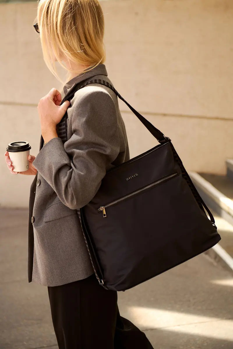 A woman in a gray blazer carries a black nylon crossbody bag with gold-tone hardware and a small silver logo on the front, while holding a takeaway coffee cup in an outdoor urban setting.