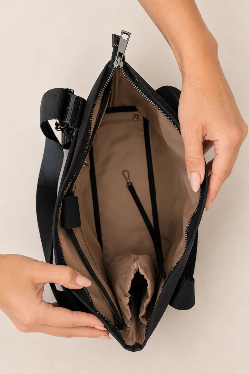 A black tote bag with a beige interior, shown open with hands holding it to display the compartments and zipper details against a light beige background.
