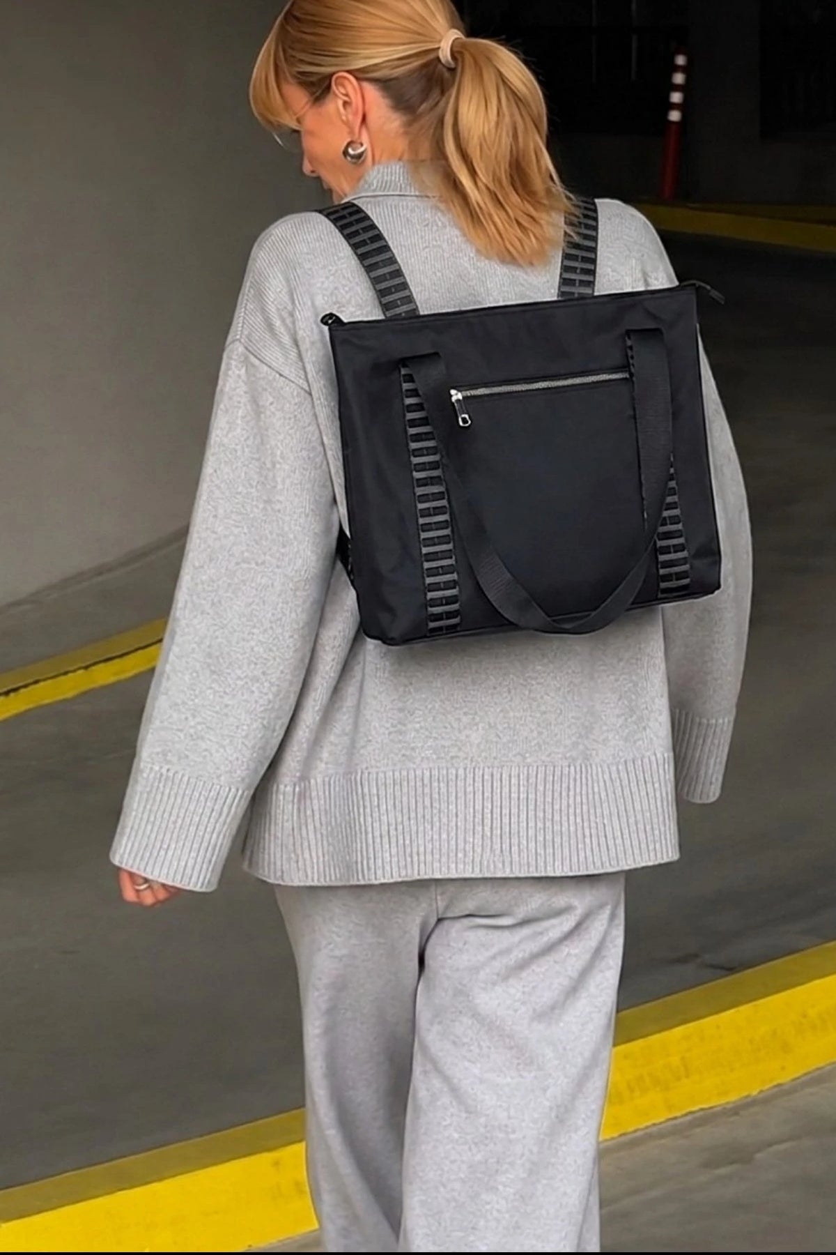A woman wearing a light gray knit sweater and matching sweatpants, shown from behind while wearing a black backpack with gray striped shoulder straps in what appears to be a concrete parking structure.