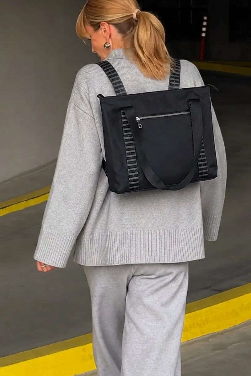 A woman wearing a light gray knit sweater and matching sweatpants, shown from behind while wearing a black backpack with gray striped shoulder straps in what appears to be a concrete parking structure.