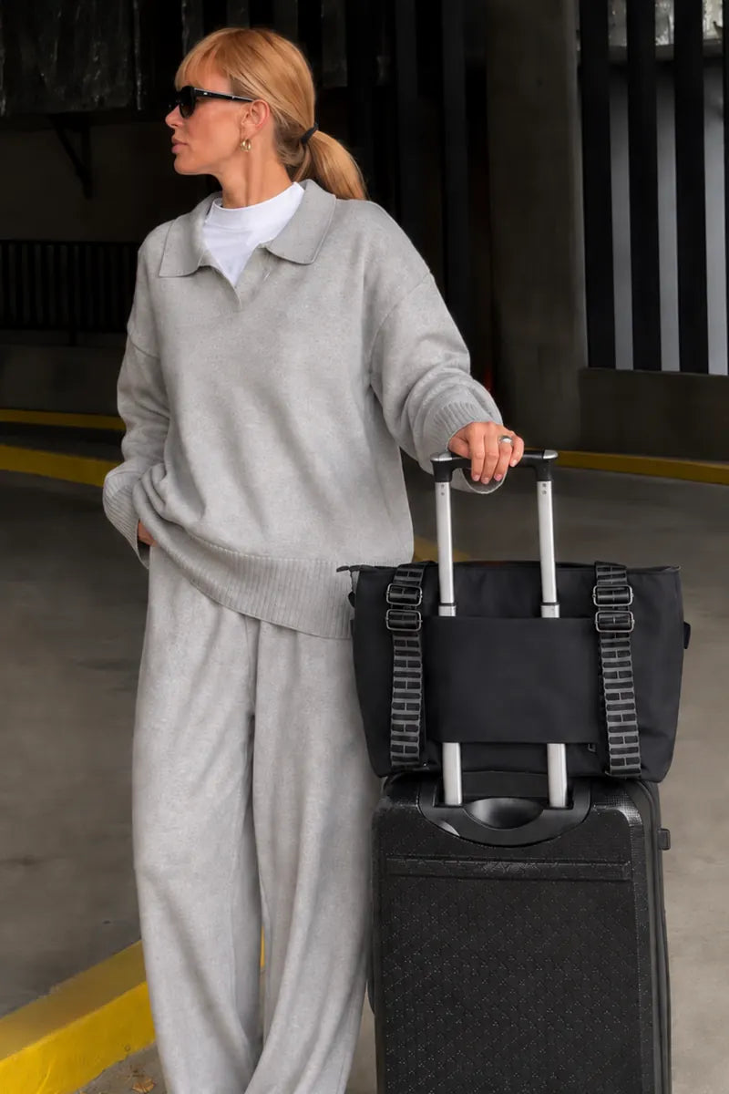A woman in a light gray knit matching set stands in a parking structure next to a black rolling suitcase, holding the handle of a black tote bag with wide straps.