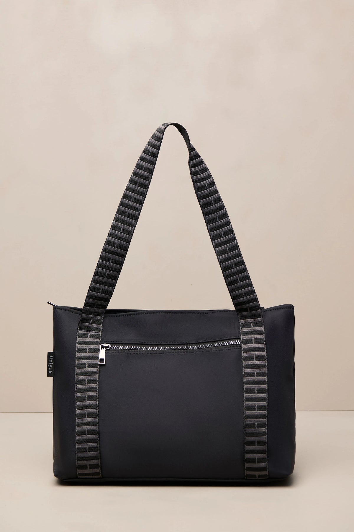 A black tote bag with textured gray and black patterned shoulder straps, shown against a neutral beige background.