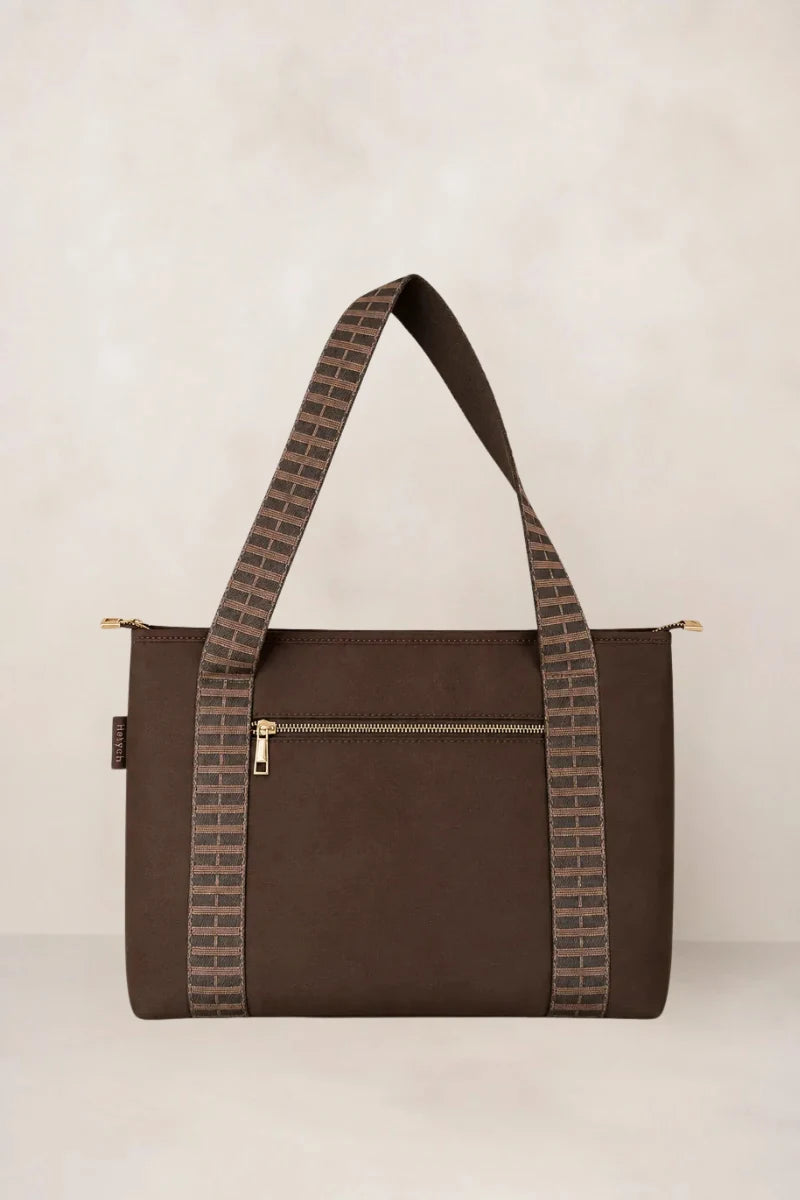 A chocolate tote bag with patterned shoulder straps, featuring a front zippered pocket and gold-tone hardware, set against a neutral beige background.