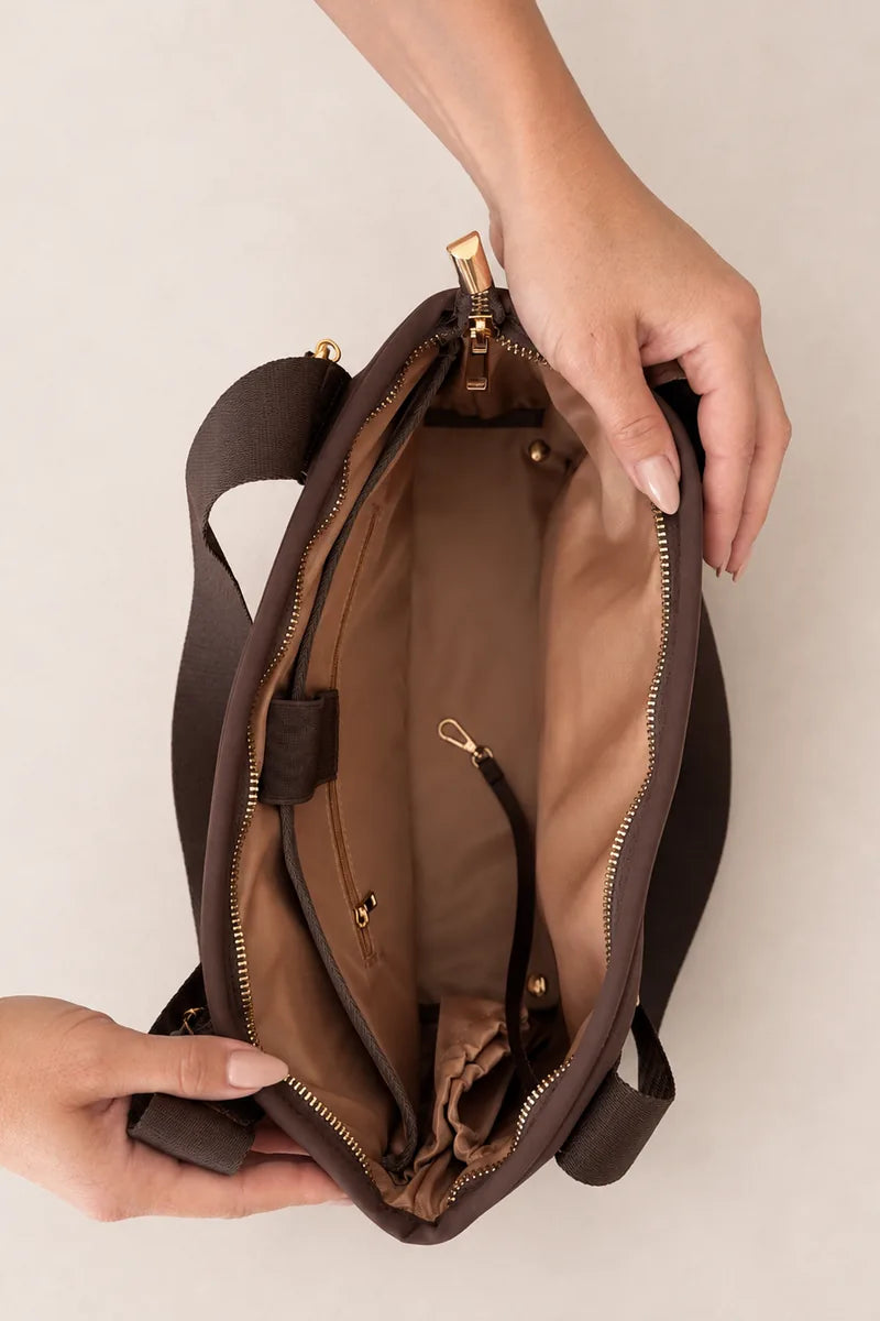 A brown chocolate bag with a gold zipper and gold hardware, shown open with hands holding it to display the interior lining and compartments against a light beige background.