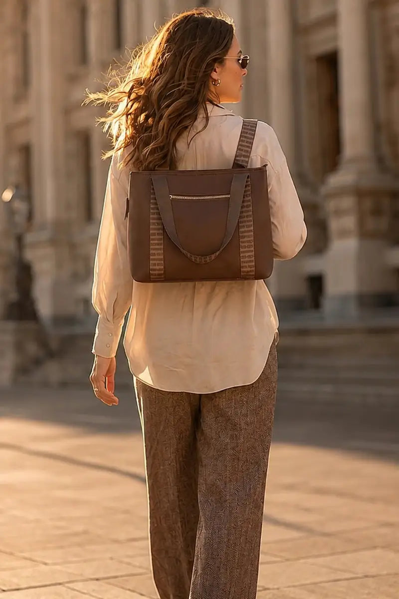 A woman seen from behind wearing a brown chocolate backpack with striped fabric side panels, styled with a cream blouse and brown trousers in an outdoor urban setting.