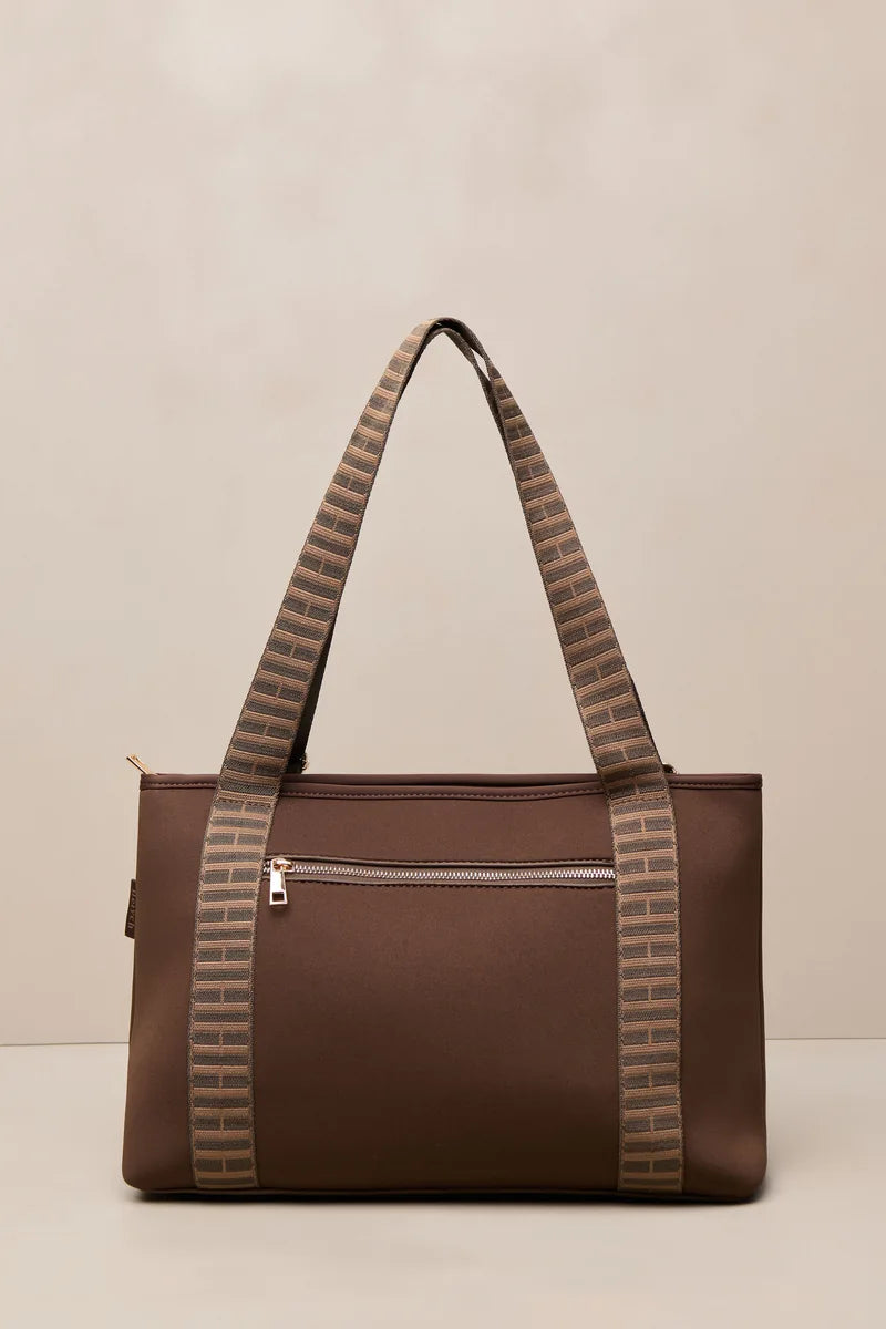 A brown chocolate tote bag with patterned shoulder straps, featuring a front zippered pocket and gold-tone hardware, set against a neutral beige background.