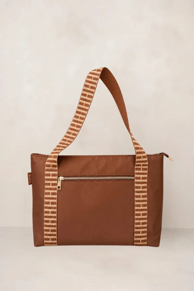 A cognac brown tote bag with a wide, patterned brown and beige shoulder strap, featuring a front zippered pocket and a small side tag, set against a neutral beige background.