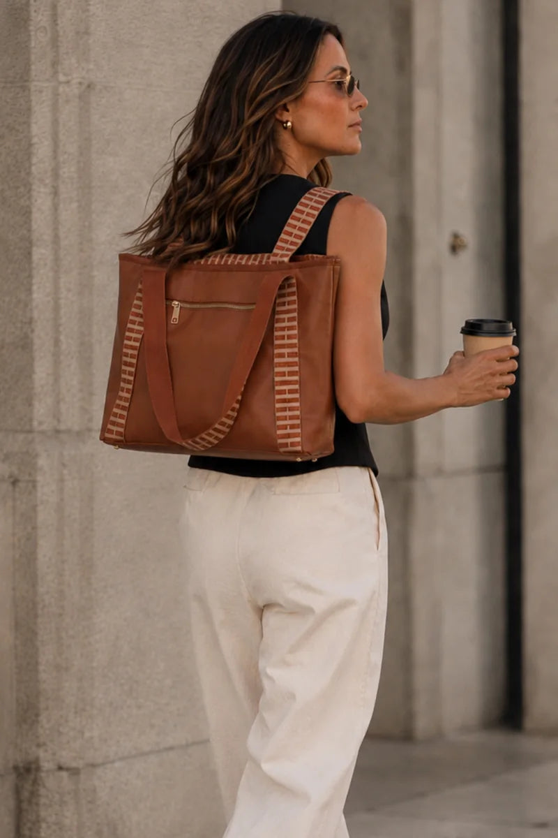 A woman wearing glasses, a sleeveless top, and cream pants carries a brown cognac tan tote bag with striped woven details on a city sidewalk next to a stone wall, holding a coffee cup.