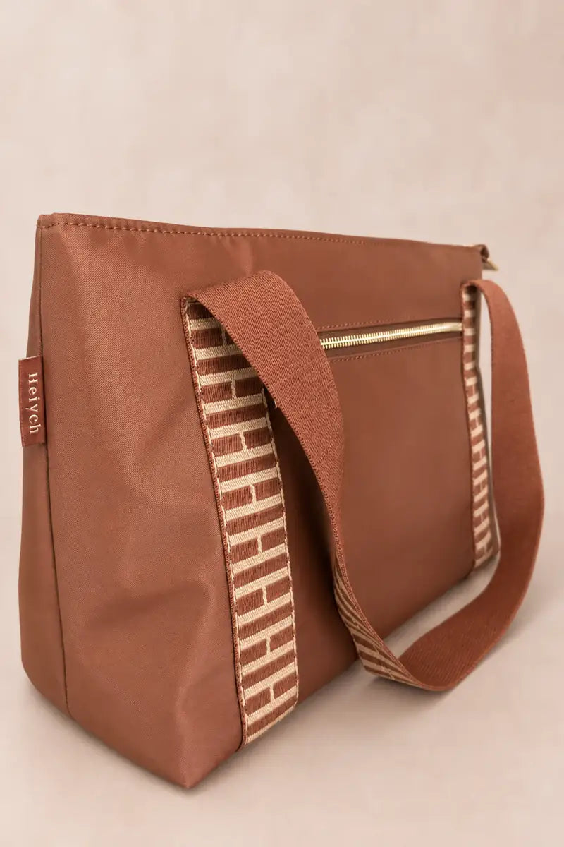 A brown cognac shoulder bag with a patterned woven strap and a small side tag reading 'HEIYCH', photographed against a plain light beige background.