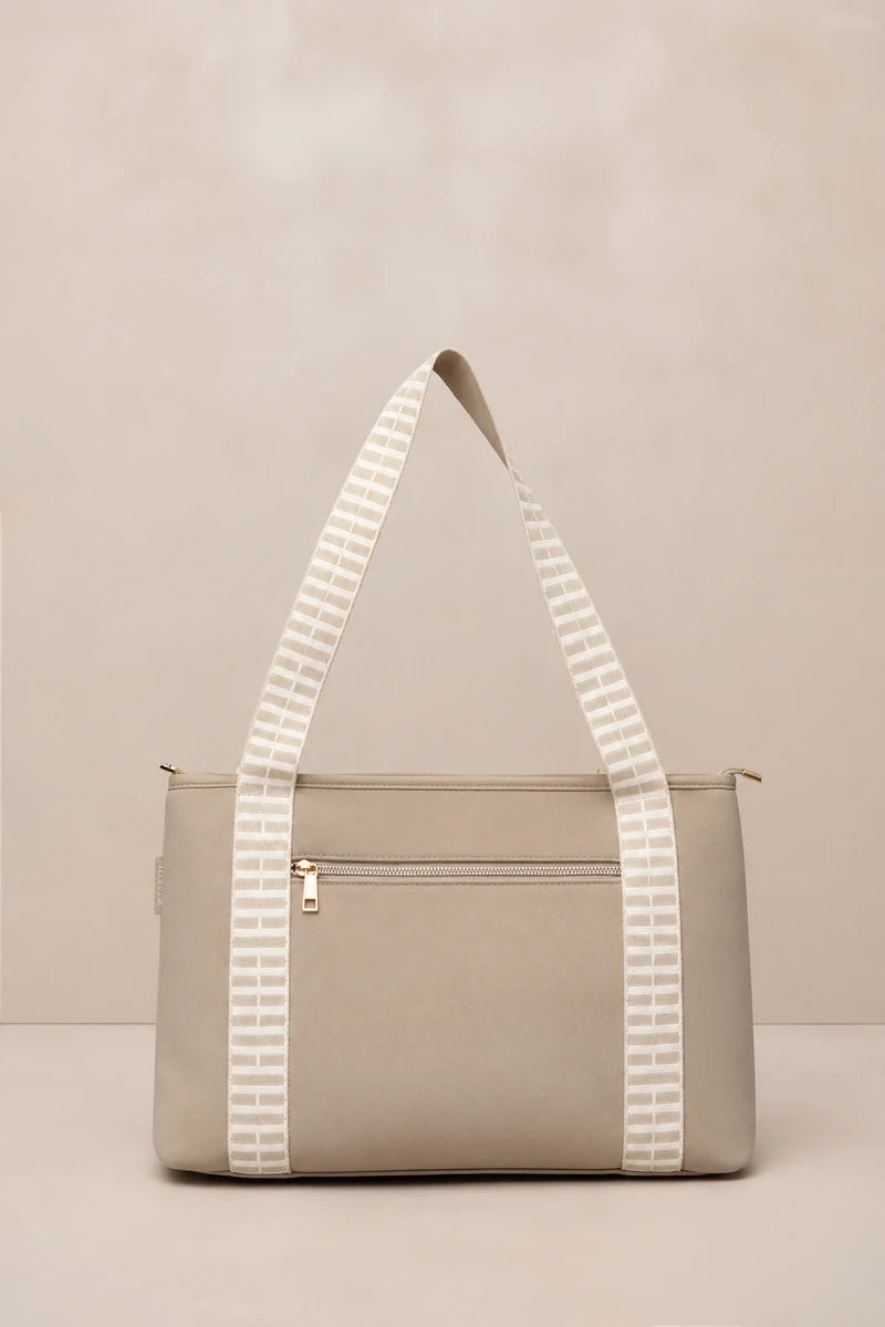 A structured taupe tote bag with a front zip pocket and dual shoulder straps featuring a white geometric pattern, set against a neutral beige background.