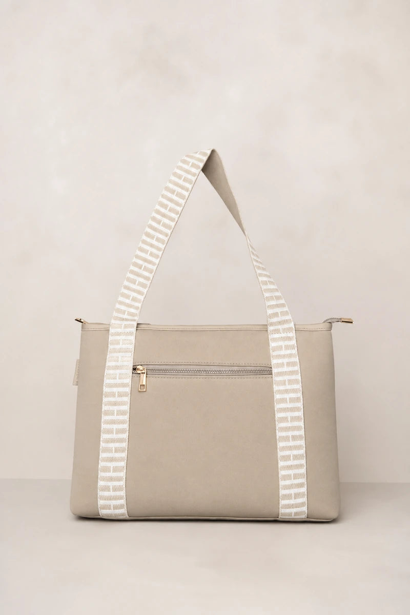 A structured taupe beige tote bag with a front zip pocket and wide shoulder straps featuring a white geometric pattern, set against a neutral beige background.