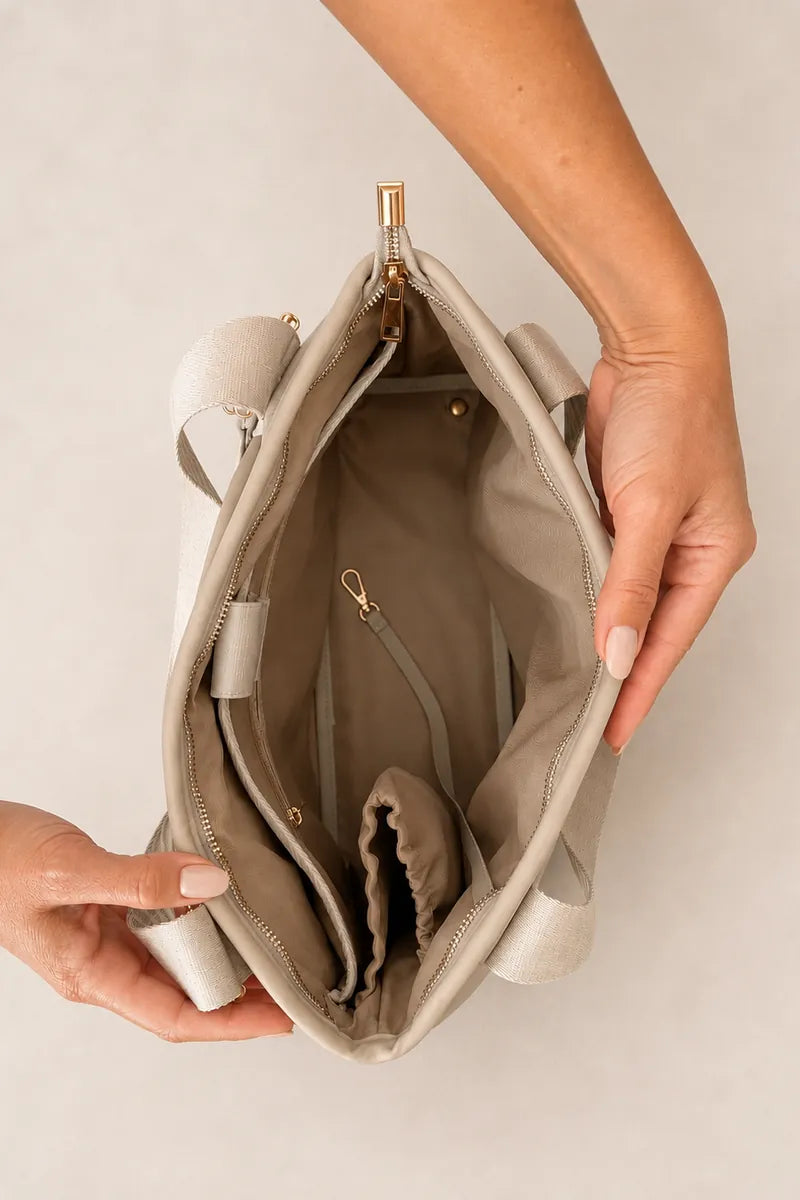 A top-down view of a taupe beige tote bag being held open by two hands to show its interior compartments and gold-tone zipper hardware, set against a neutral light gray background.