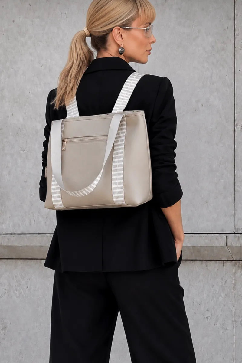 A woman wearing a blazer and pants is shown from the back, carrying a structured taupe beige tote bag with white striped shoulder straps against a grey concrete wall.